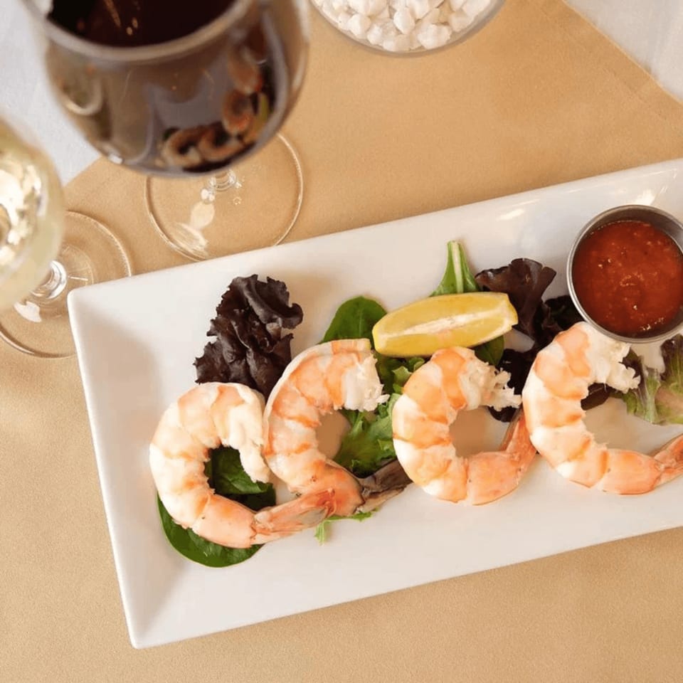 Shrimp cocktail with lemon, greens, and sauce on a white plate, with wine glasses nearby.